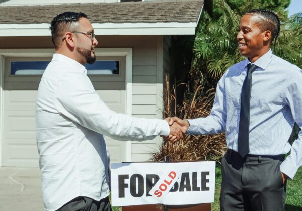 Two men shaking hands in front of house sold sign, sealing real estate deal outdoors.