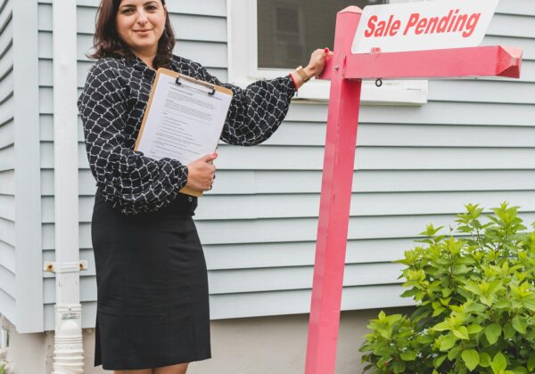 Confident female realtor standing next to a Sale Pending sign outside a house, showcasing success.